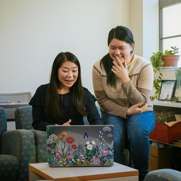 Two students look at a laptop with stickers of flowers and laugh at what they see.